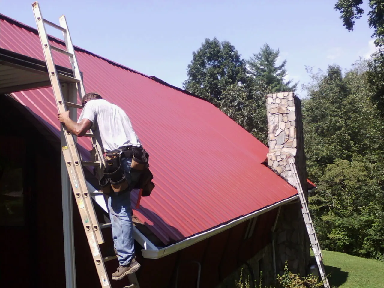 Roofer on a ladder beside a red metal roof and stone chimney — Soffit & Fascia Repair by Roofing Brothers in Humphrey